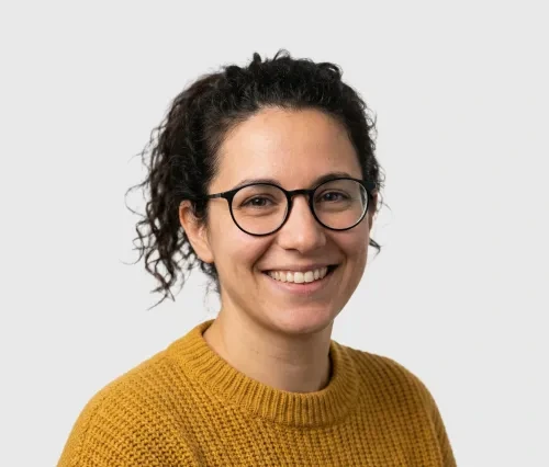 Smiling woman with curly dark hair, glasses, and a mustard yellow knit sweater against a gray background.