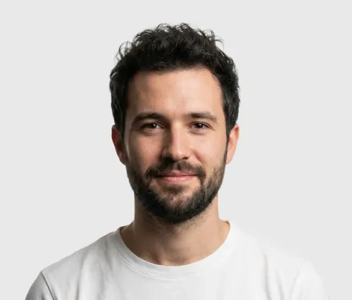 Smiling man with curly dark hair and beard wearing a white T-shirt against gray background.