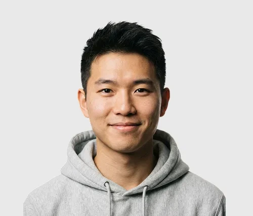 Young man with short black hair smiling in a light gray hoodie against white background.
