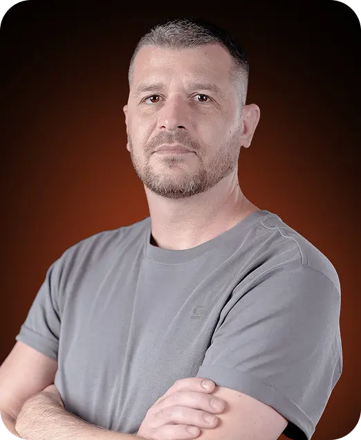 Portrait of a man with short hair and beard wearing a gray t-shirt, standing with arms crossed against a dark orange background.