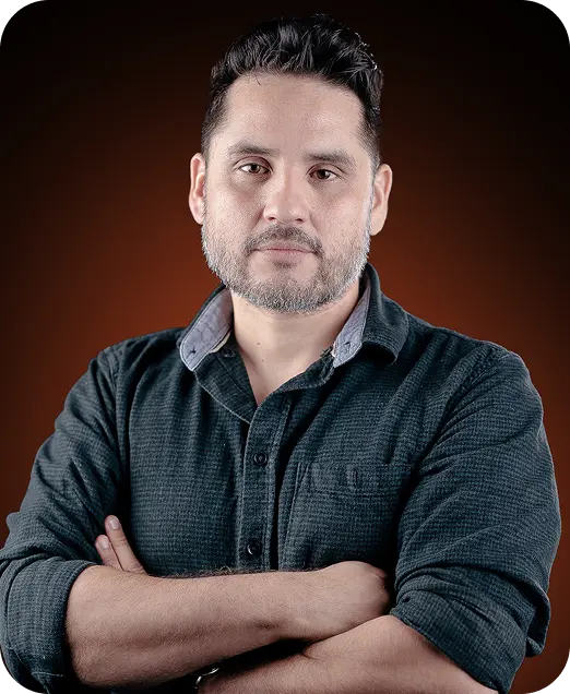 Portrait of a middle-aged man with short dark hair and a beard, wearing a dark button-up shirt with arms crossed against a brown background.