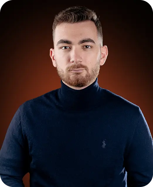Portrait of a man with short dark hair and a beard wearing a navy blue turtleneck sweater against a dark brown background.