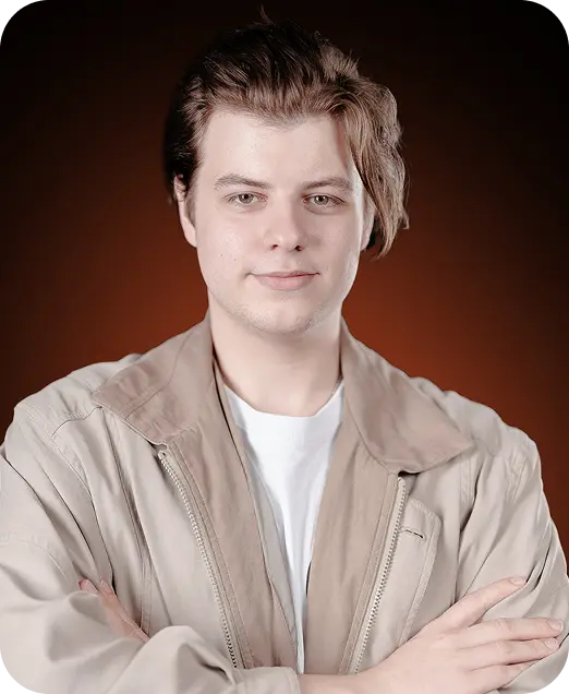 Young man with light skin and brown hair wearing a beige jacket over a white shirt, posing with arms crossed against a dark orange background.