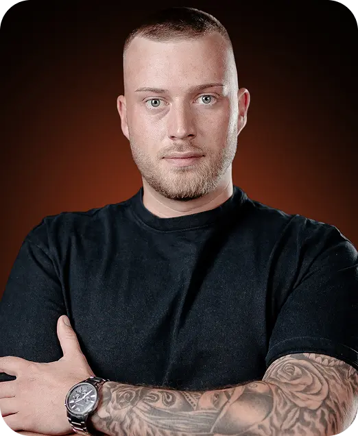 Portrait of a man with close-cropped hair and a beard wearing a black t-shirt, showing tattooed arms and a wristwatch, against a dark brown background.