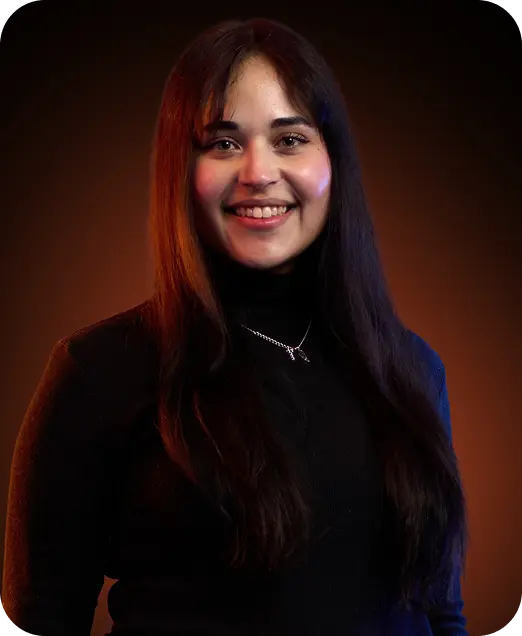 Smiling young woman with long dark hair wearing a black turtleneck and a silver necklace against a dark blurred background.