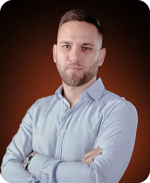 Confident young man with short beard and hair wearing a striped button-up shirt standing with arms crossed against a dark brown background.