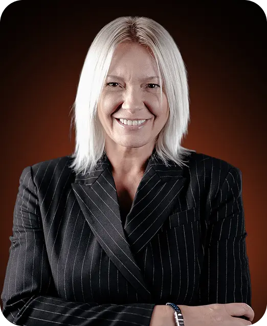 Smiling middle-aged woman with straight blonde hair wearing a black pinstripe blazer against a dark background.