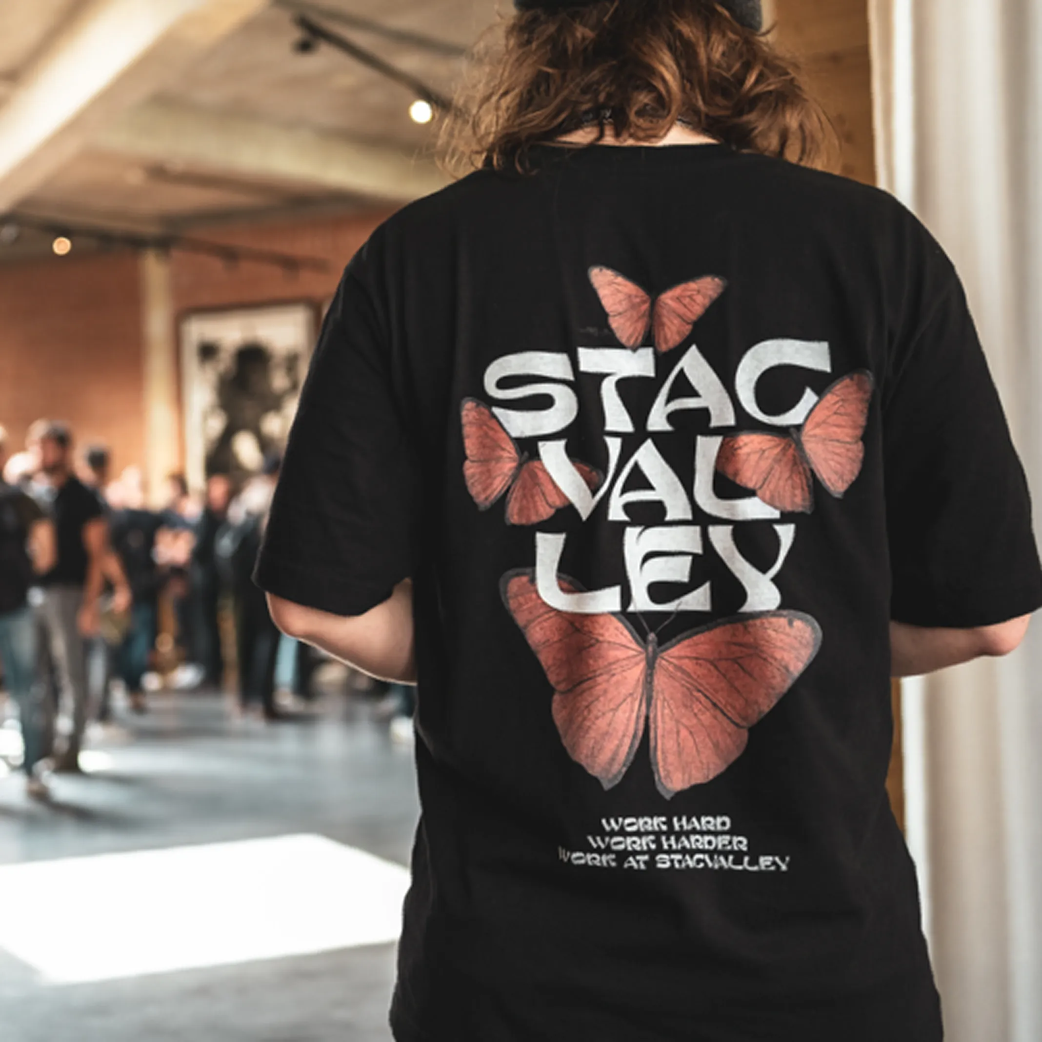 Person wearing a black Stagvalley t-shirt with orange butterflies and text reading 'Work Hard Work Harder Work At Stagvalley' on the back in a busy indoor setting.