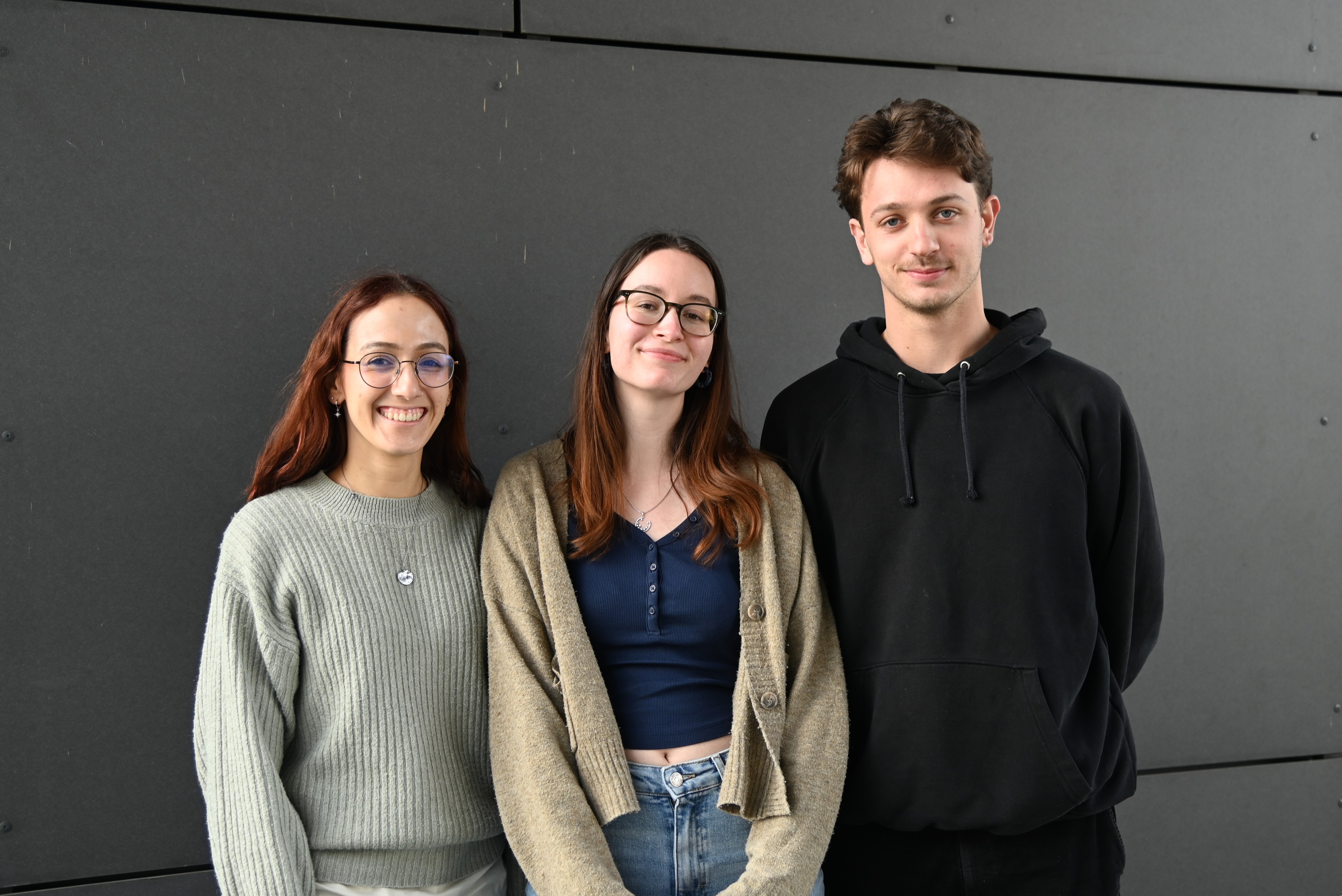 Three young adults standing side by side smiling against a dark wall background.