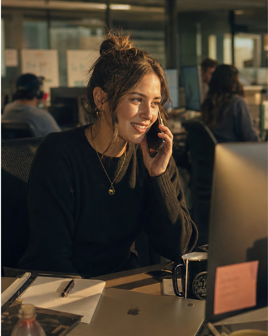 Smiling woman talking on phone while working at a desk with computer and notebook in office.