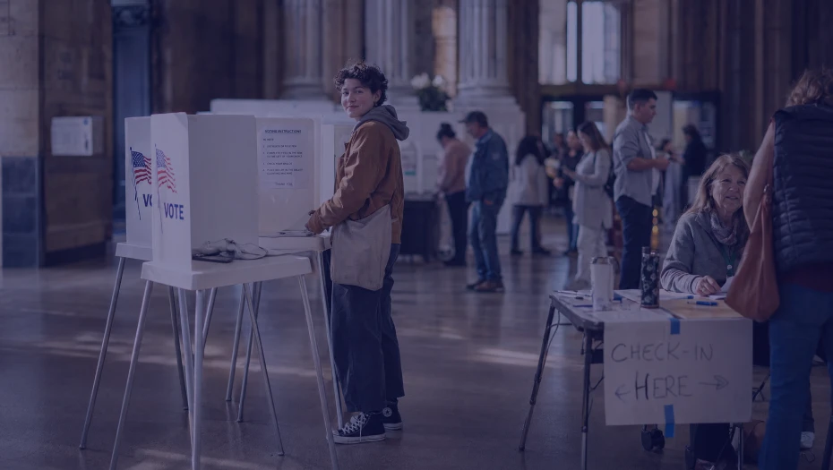 Person casting a ballot at a polling station with check-in table and other voters in the background.