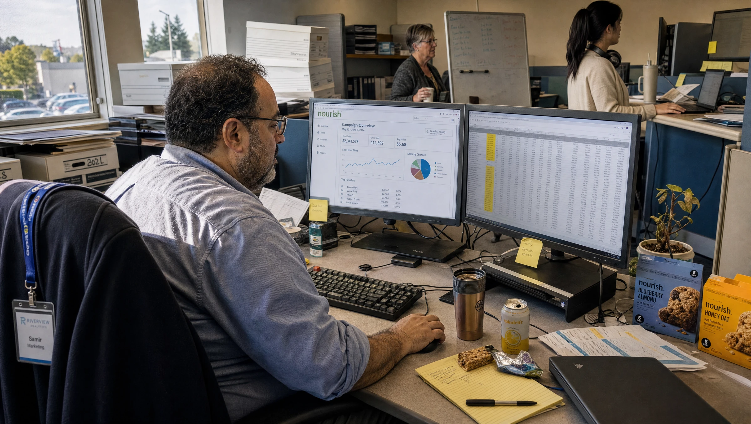 A marketing analyst cross-checking a campaign dashboard against a sales spreadsheet on two monitors in an open-plan office