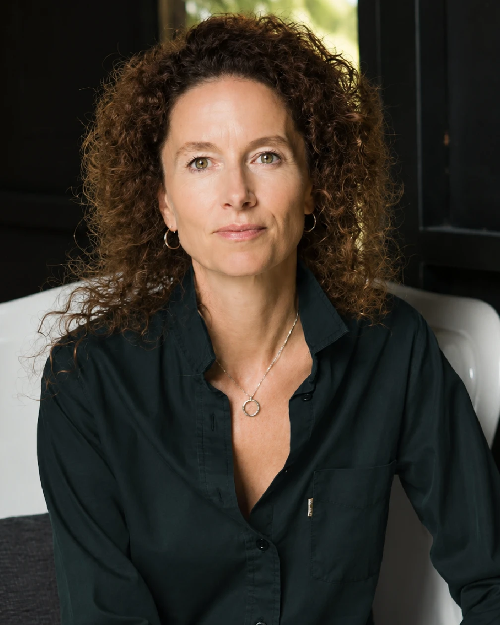 Woman with curly hair wearing a dark green button-up shirt and gold hoop earrings looking at camera.