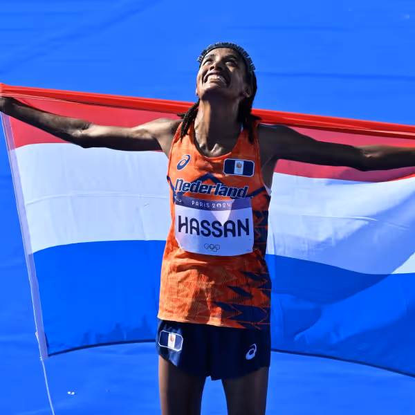 Sifan Hassan wearing an orange Netherlands uniform holding a Dutch flag behind her, looking up and smiling on a blue track during her Paris 2024 Marathon win.