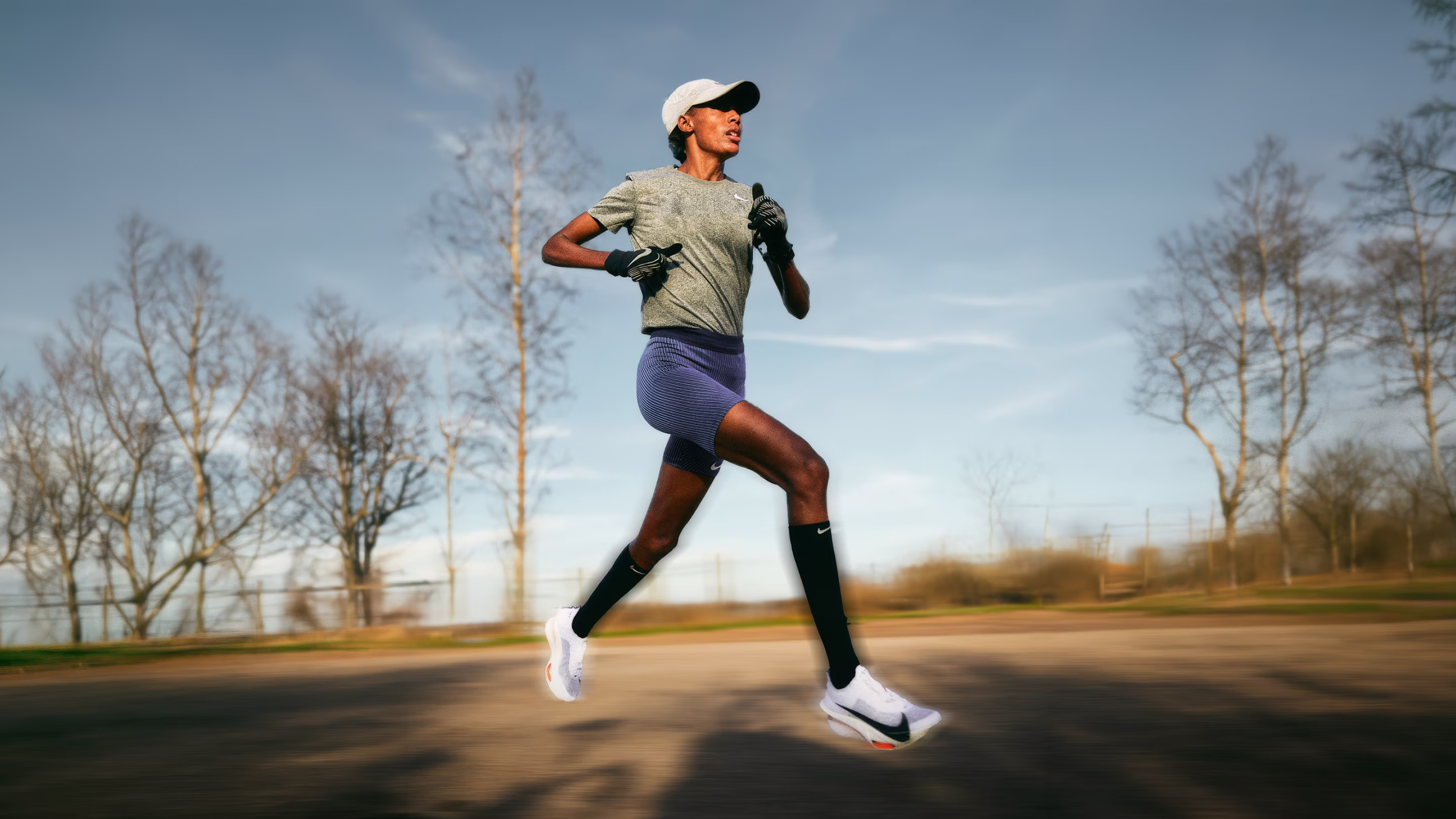 Sifan Hassan in athletic gear sprinting outdoors on a paved path with bare trees and a blue sky background.