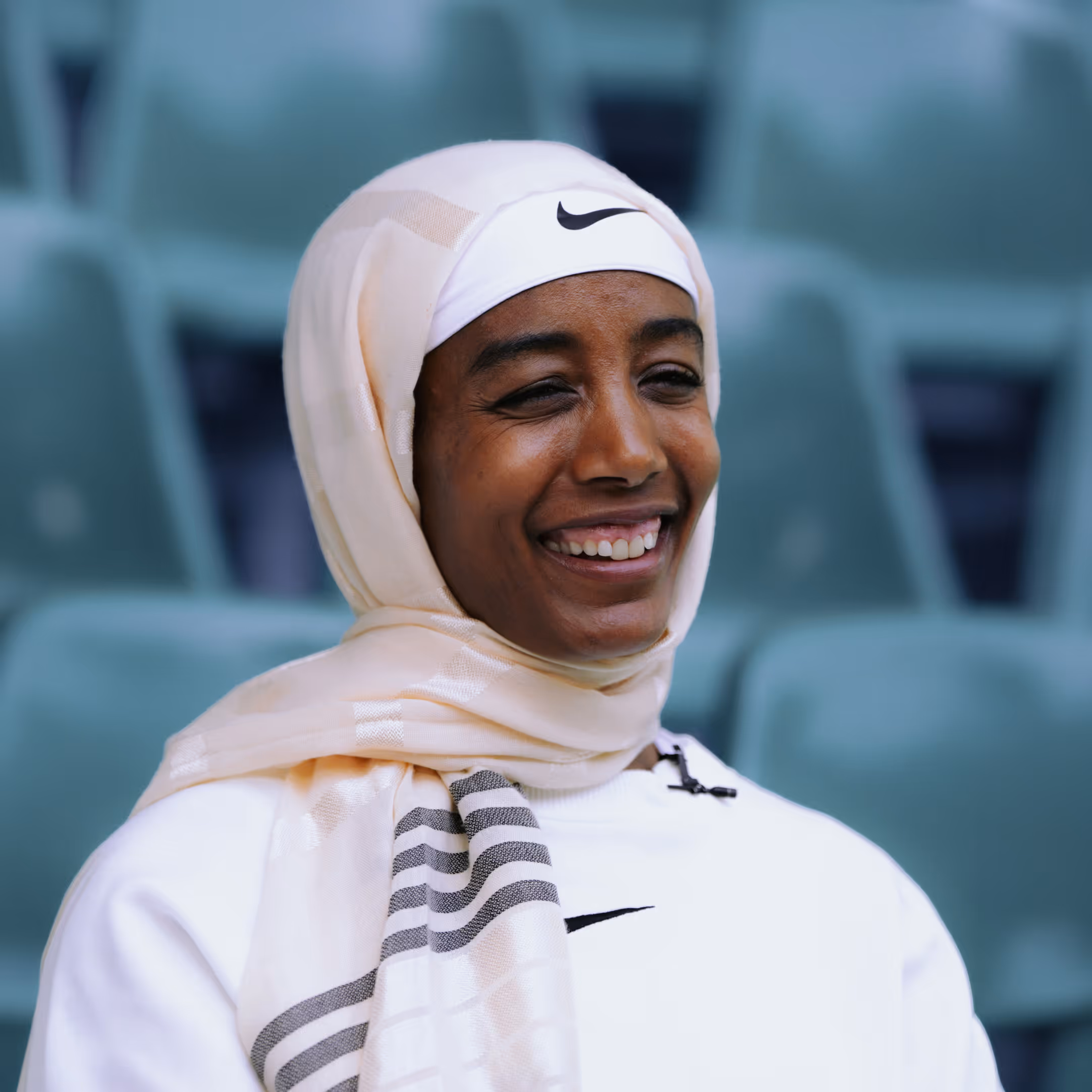 Smiling Sifan Hassan wearing a light beige hijab and white Nike headband and sweatshirt, seated in an outdoor stadium.
