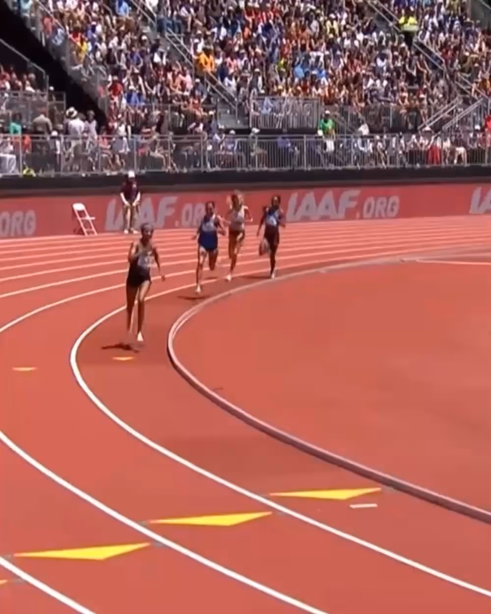 Four female athletes competing in a 3000m race on an outdoor track with a crowd watching from the stands.