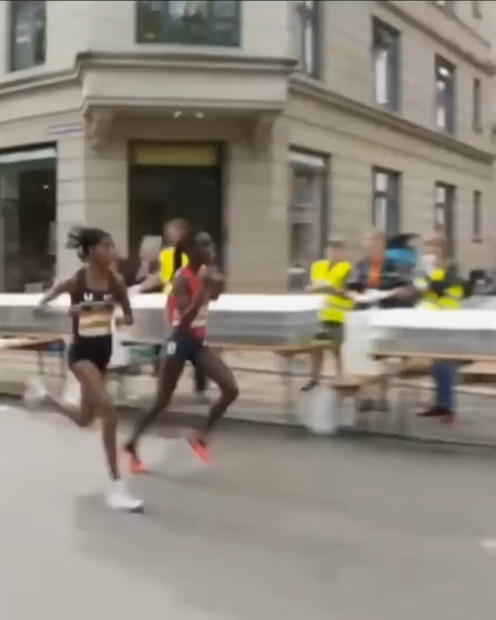 Two female runners competing in a street race with volunteers in yellow vests in the background.