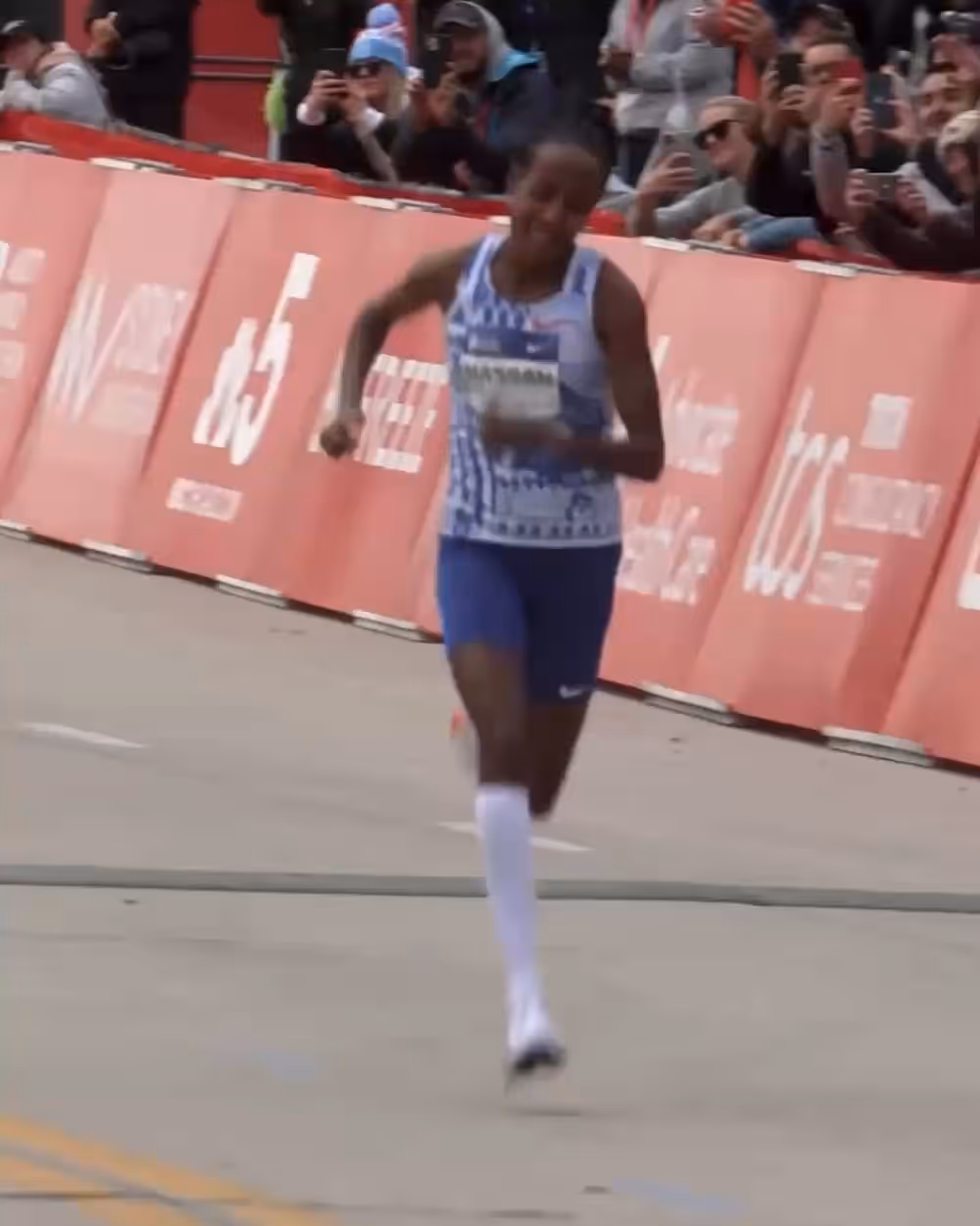 Marathon runner in blue and white gear nearing the finish line with crowd cheering behind red barriers.