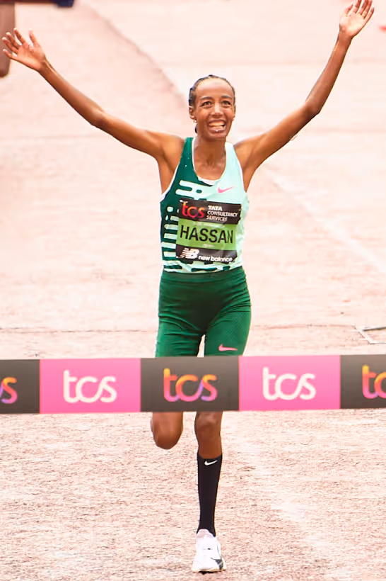 Female marathon runner Hassan crossing the finish line with arms raised in victory at the London Marathon.