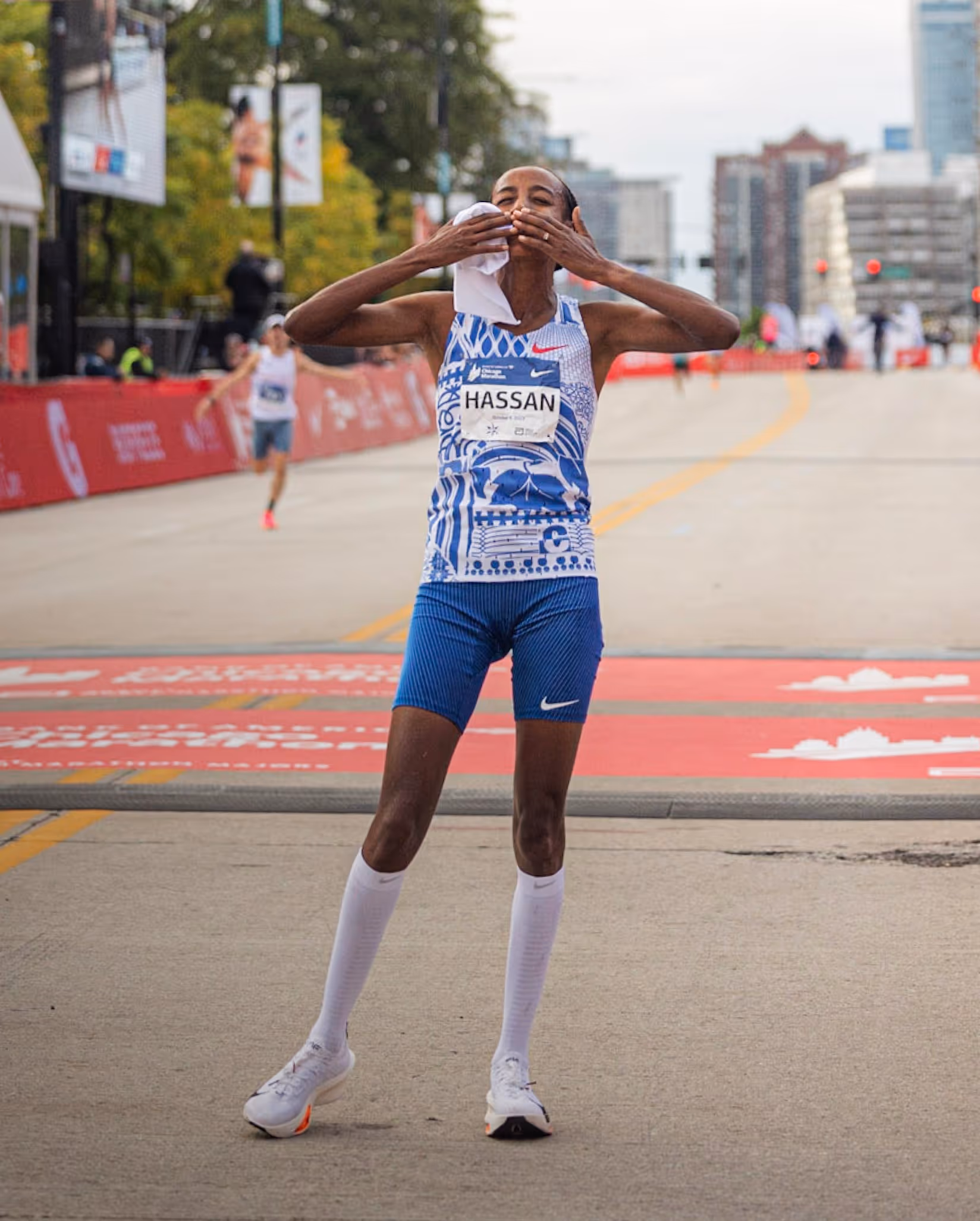 Runner wearing a blue and white outfit with 'HASSAN' on the bib crosses the marathon finish line, blowing kisses and holding a towel.