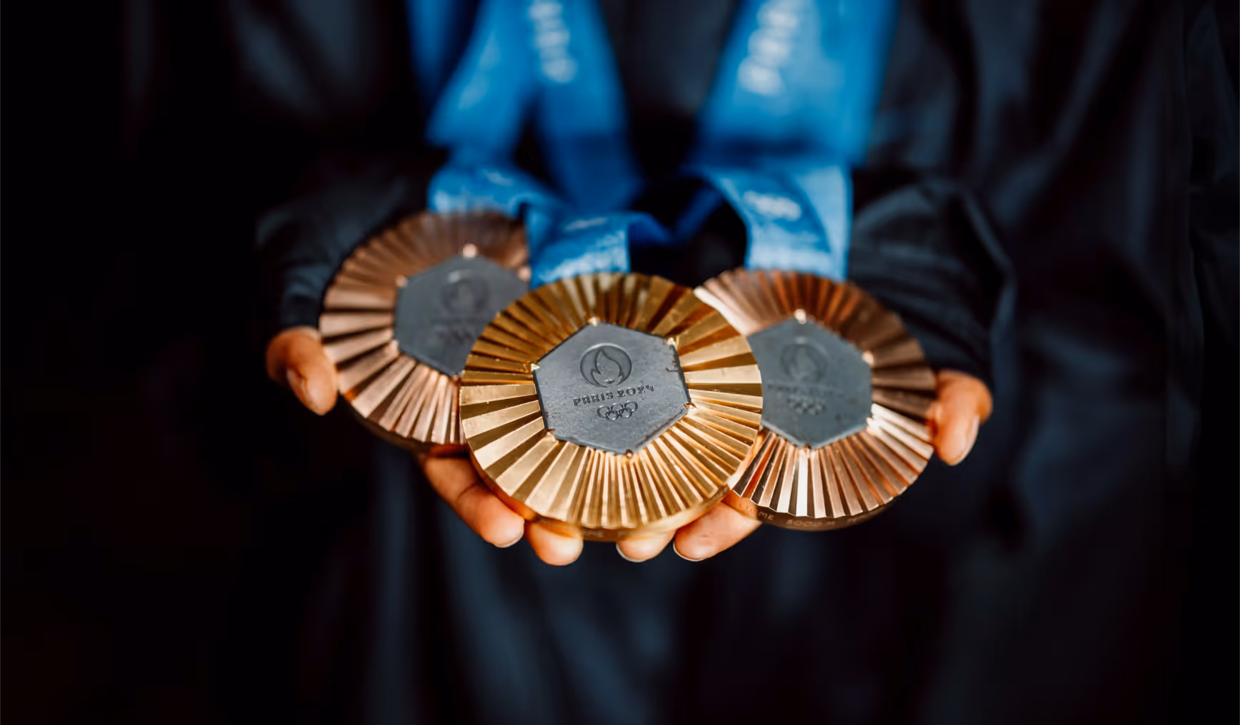 Hands holding three Olympic medals for Paris 2024 with blue ribbons, including gold, silver, and bronze.