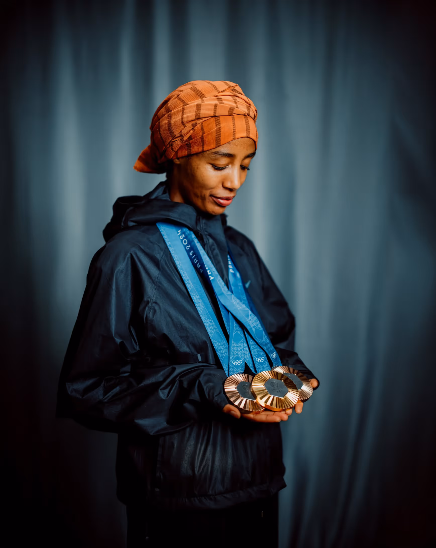 Woman wearing an orange headscarf and black jacket looking down at three medals with blue ribbons in her hands.