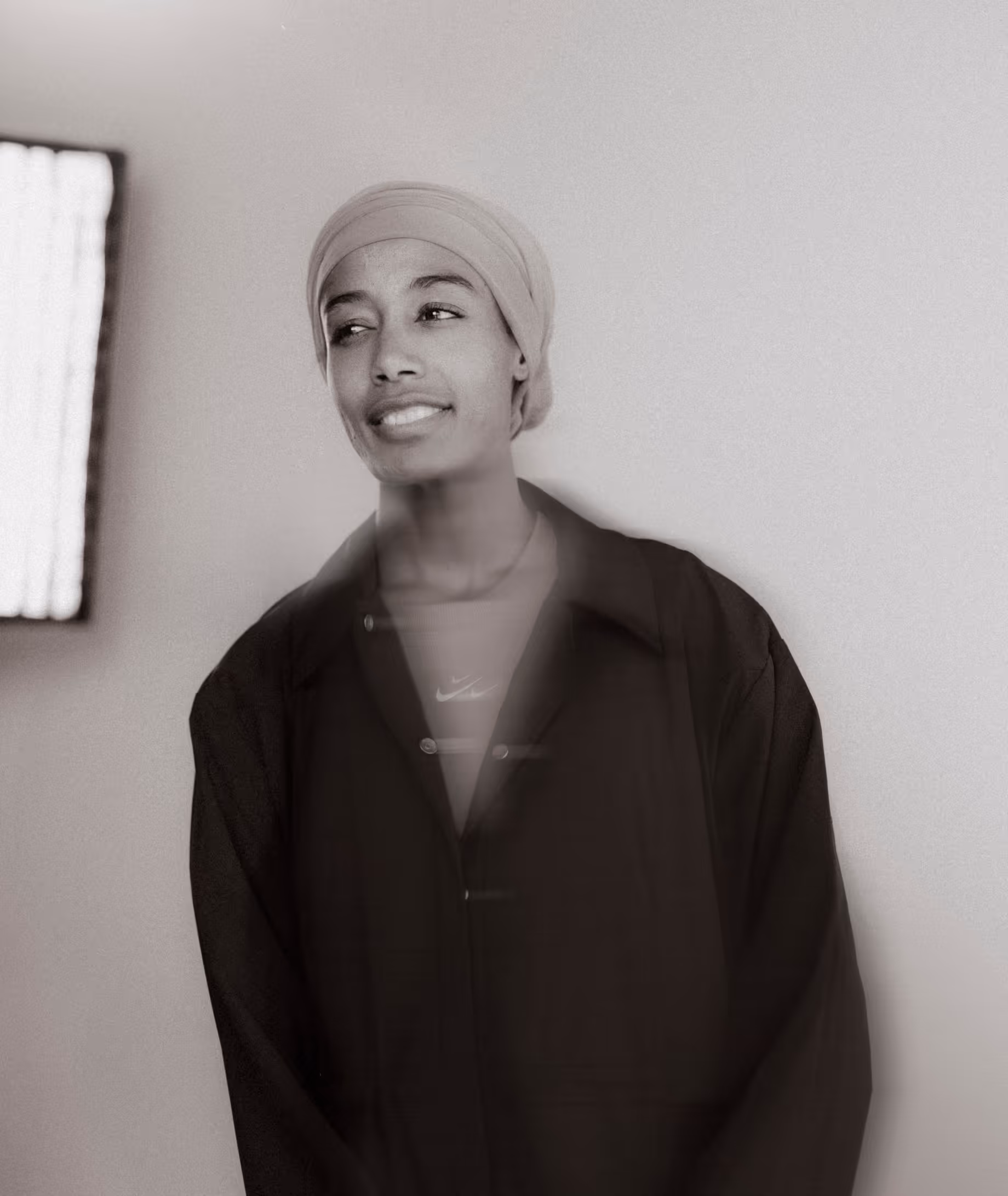 Black and white portrait of a smiling person wearing a headwrap and dark jacket against a plain wall.
