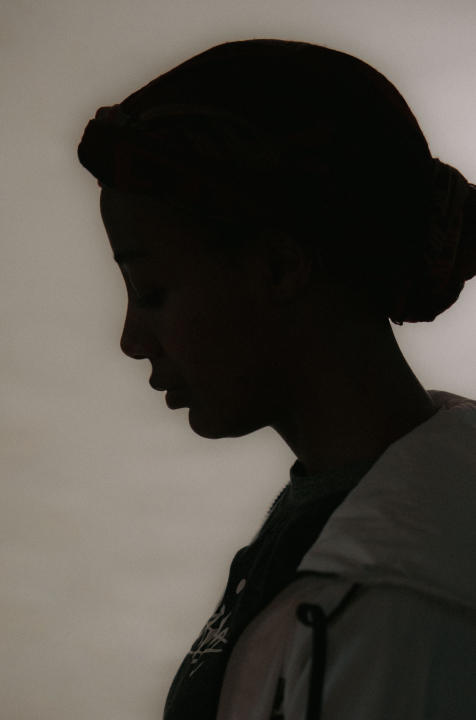 Silhouette of a person in profile wearing a headwrap and a light jacket against a plain background.