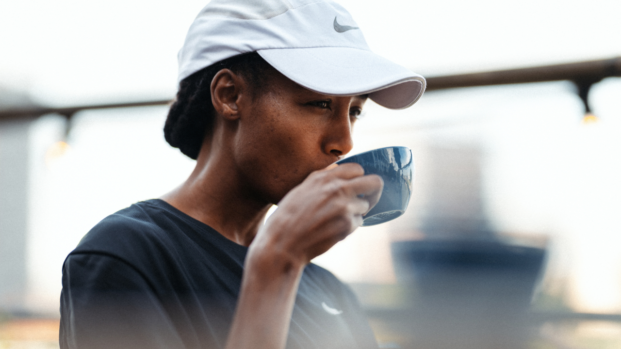 Woman in a white Nike cap and black shirt drinking from a blue cup outdoors.