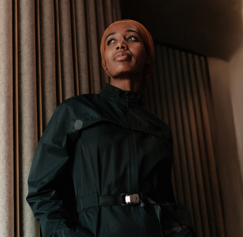 Confident woman wearing a black jacket and an orange headscarf standing indoors near curtains.