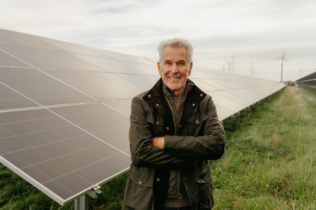 Älterer Mann steht lächelnd mit verschränkten Armen vor Sonnenkollektoren in einer grünen Wiese mit Windrädern im Hintergrund.