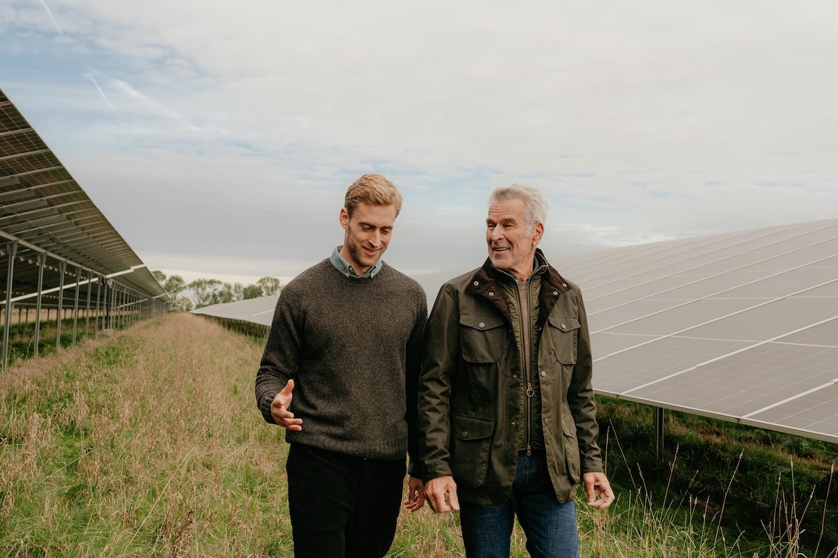 Zwei Männer gehen und sprechen neben großen Solarpaneelen in einem Feld unter bewölktem Himmel.
