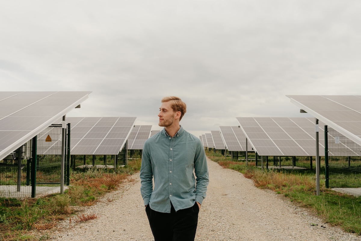 Mann in grünem Hemd geht zwischen Reihen von Solarpanelen auf einem Kiesweg bei bewölktem Himmel.