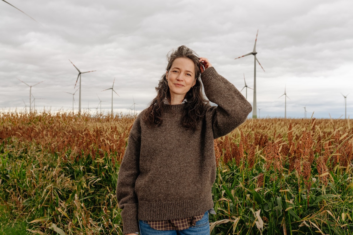 Frau mit braunem Pullover steht in einem Feld mit Windrädern im Hintergrund bei bewölktem Himmel.