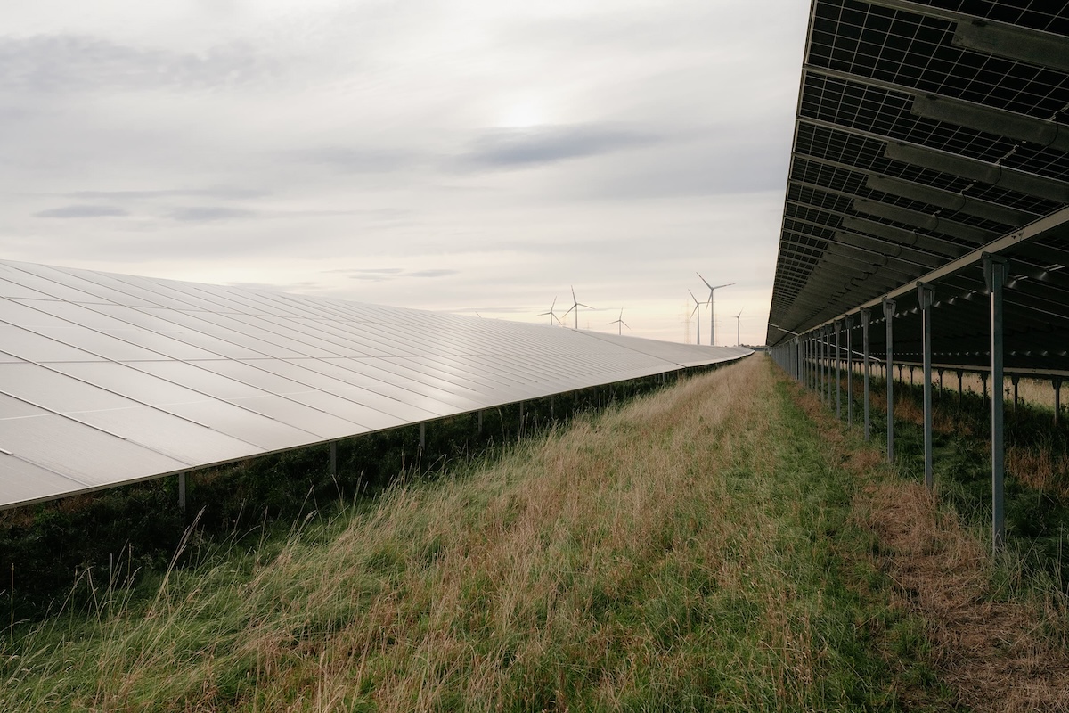 Sonnenkollektoren in einem Feld mit Windkraftanlagen im Hintergrund unter bewölktem Himmel.