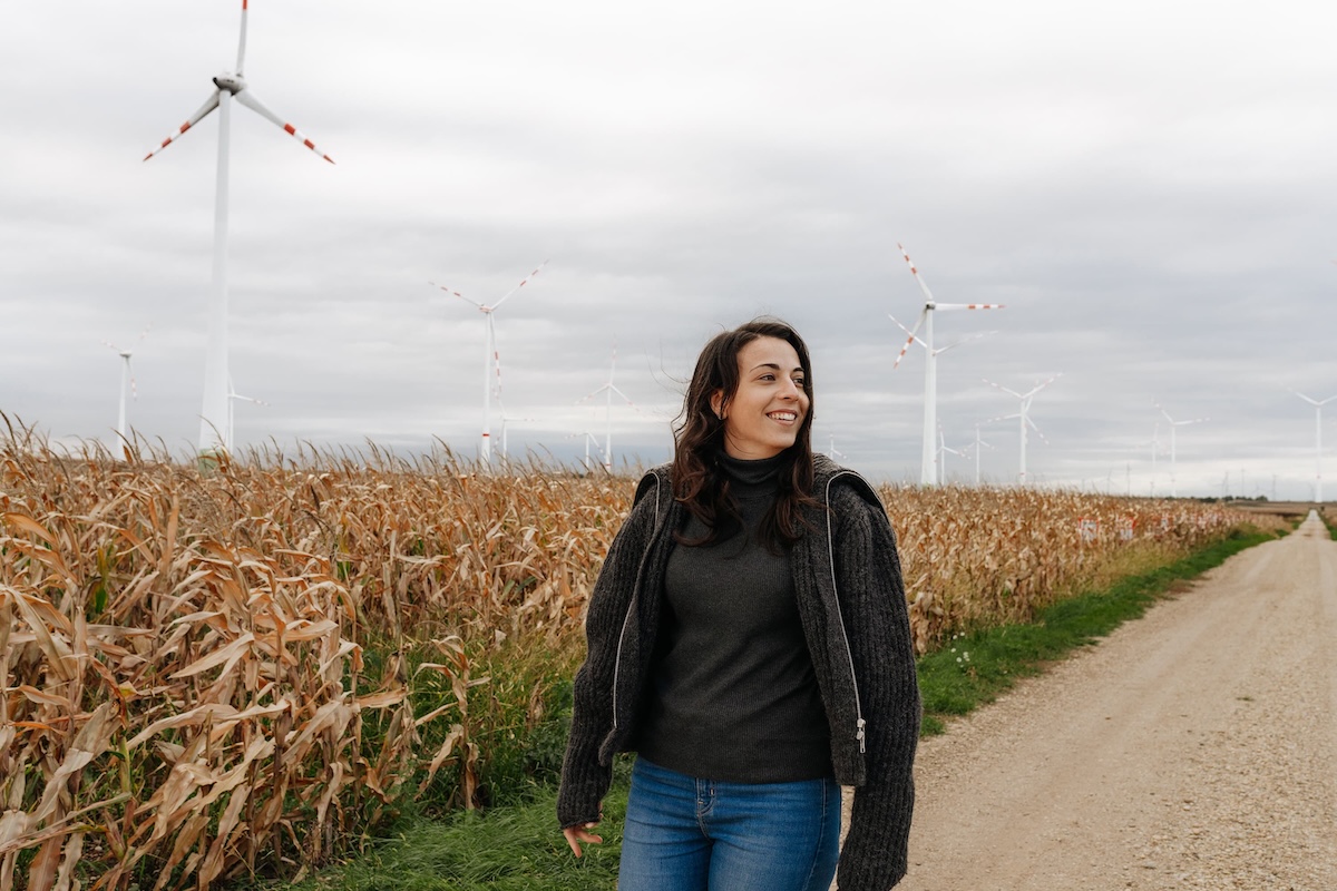 Frau in dunklem Pullover und Jeans geht auf einem Feldweg neben einem Maisfeld mit Windrädern im Hintergrund unter bewölktem Himmel.