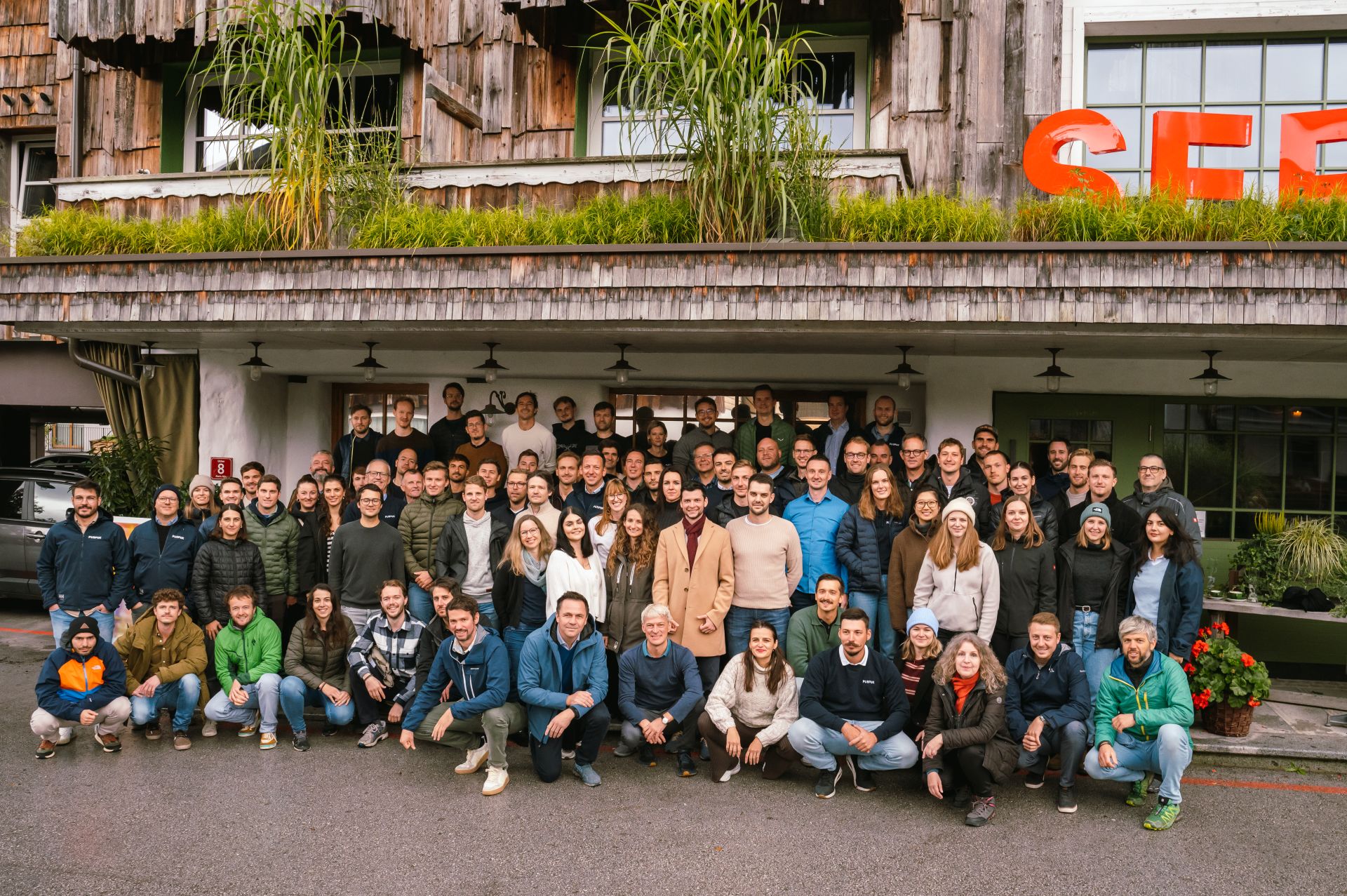 Großes Teamfoto von etwa 70 Personen, die draußen vor einem Holzgebäude mit grünen Fensterläden und Pflanzen in der Nähe eines rot-orangefarbenen Schriftzugs posieren. Die Gruppe ist gemischt geschlechtlich und in mehreren Reihen angeordnet, vorne hocken einige, während andere stehen. Alle tragen lässige Kleidung, viele mit Jacken und Pullovern.