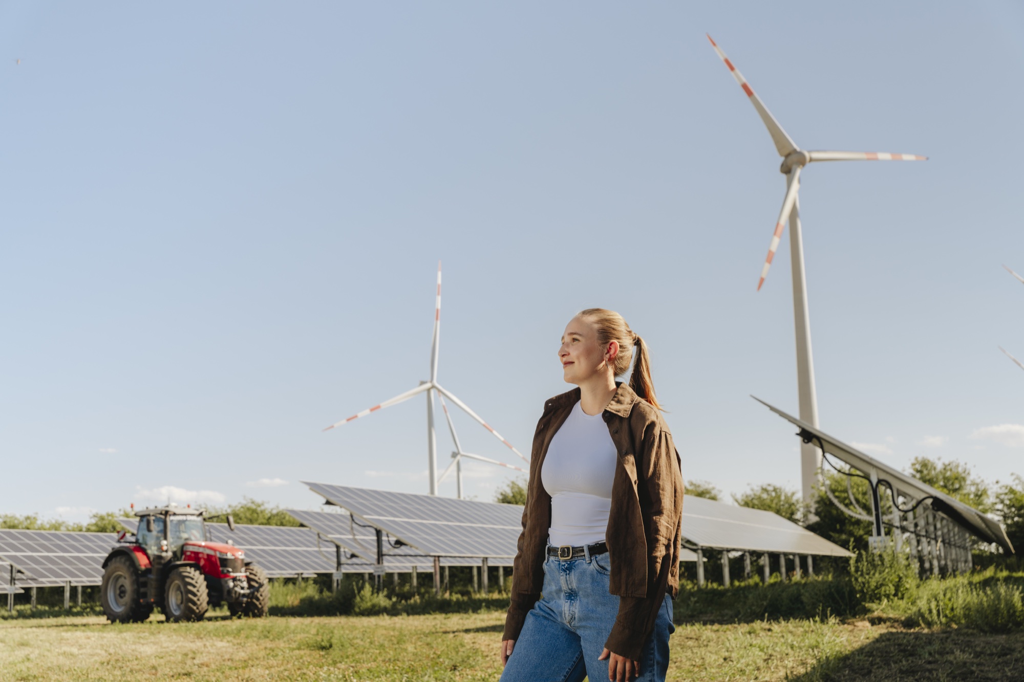 Frau steht vor Solarpanelen, Windrädern und rotem Traktor auf grünem Feld bei blauem Himmel.