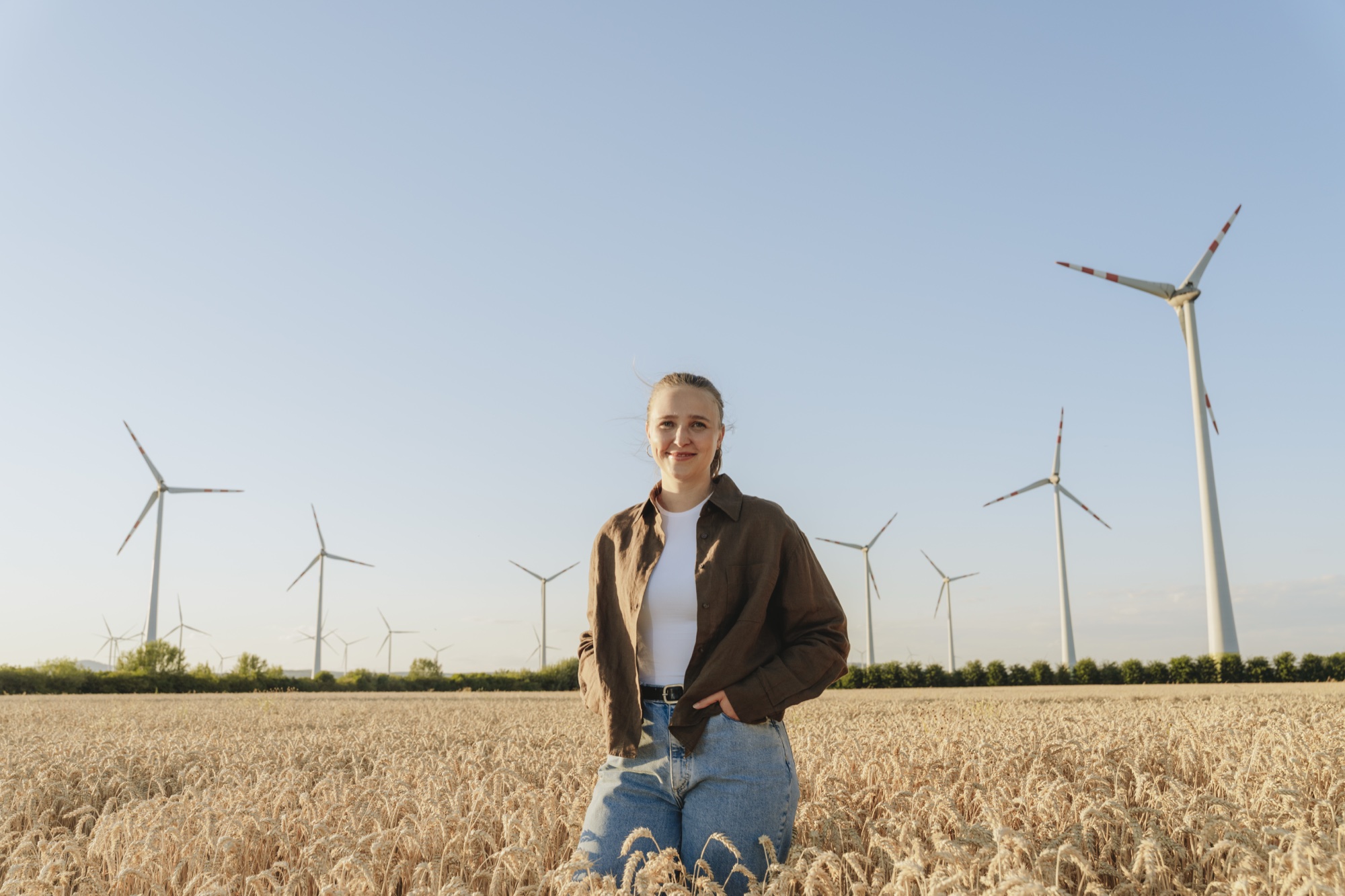 Junge Frau steht in einem Weizenfeld mit Windkraftanlagen im Hintergrund bei klarem Himmel.