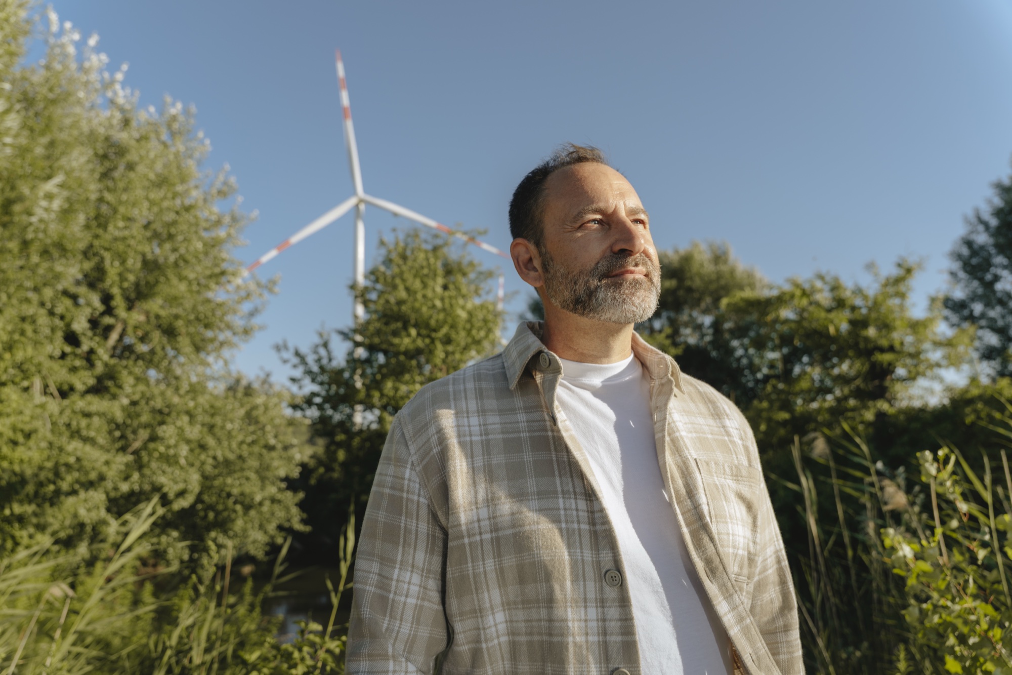 Mann in kariertem Hemd steht im Grünen vor Windkraftanlage und klarem Himmel.