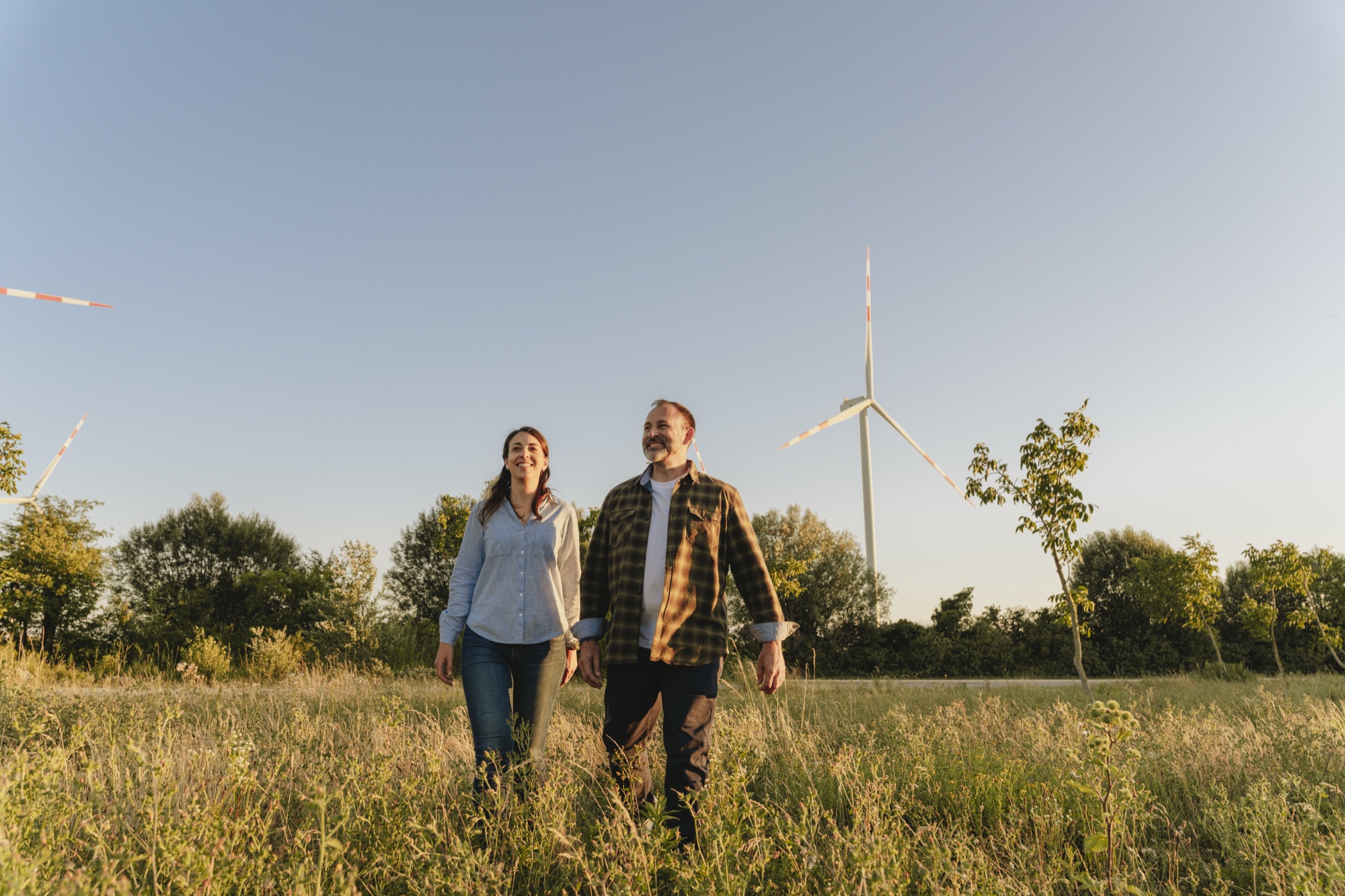 Ein Mann und eine Frau gehen lachend über eine Wiese mit Windrädern im Hintergrund bei Sonnenuntergang.