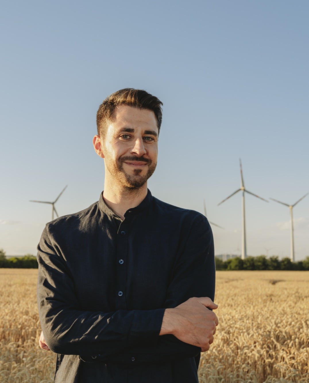 Mann in dunklem Hemd mit verschränkten Armen steht vor Feld mit Windrädern im Hintergrund.