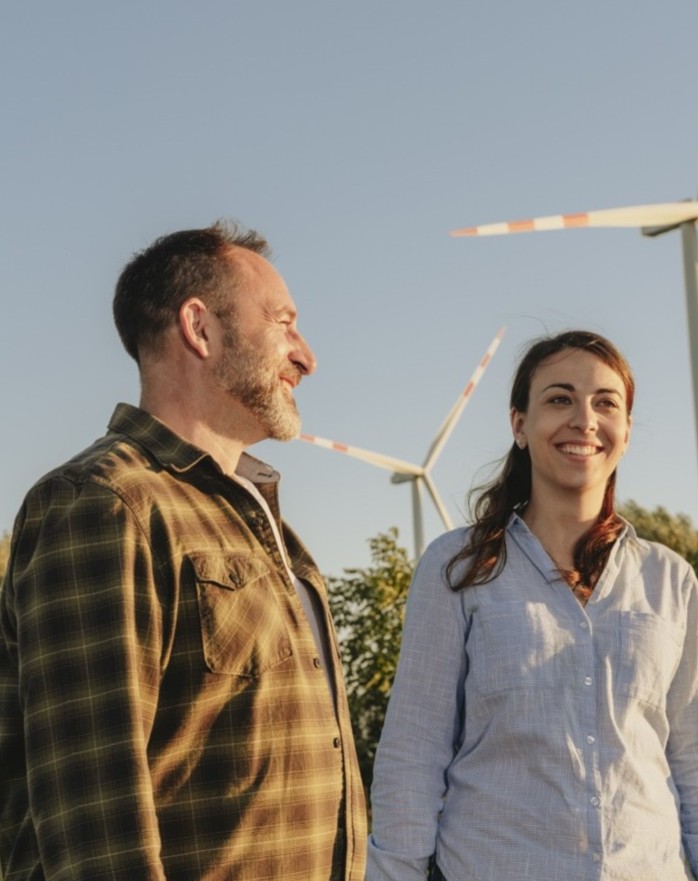 Mann und Frau stehen vor Windrädern und lächeln bei klarem Himmel.