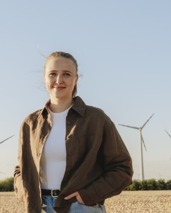 Lächelnde Frau mit brauner Hemdbluse und weißem T-Shirt vor Windrädern und blauem Himmel.
