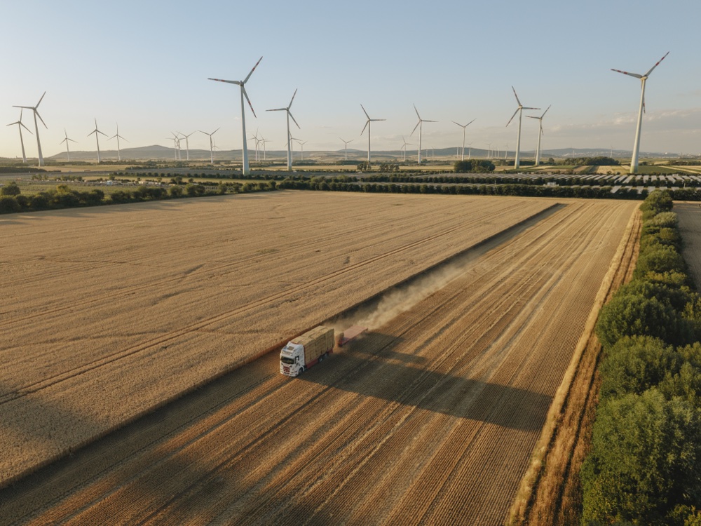 Ein landwirtschaftliches Feld mit einer Erntemaschine, die Stroh oder Getreide sammelt, vor einer Reihe von Windkraftanlagen unter klarem Himmel.