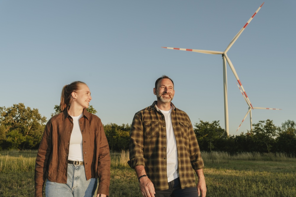 Ein Mann und eine Frau gehen lächelnd auf einer grünen Wiese mit Windkraftanlagen im Hintergrund bei klarem Himmel.