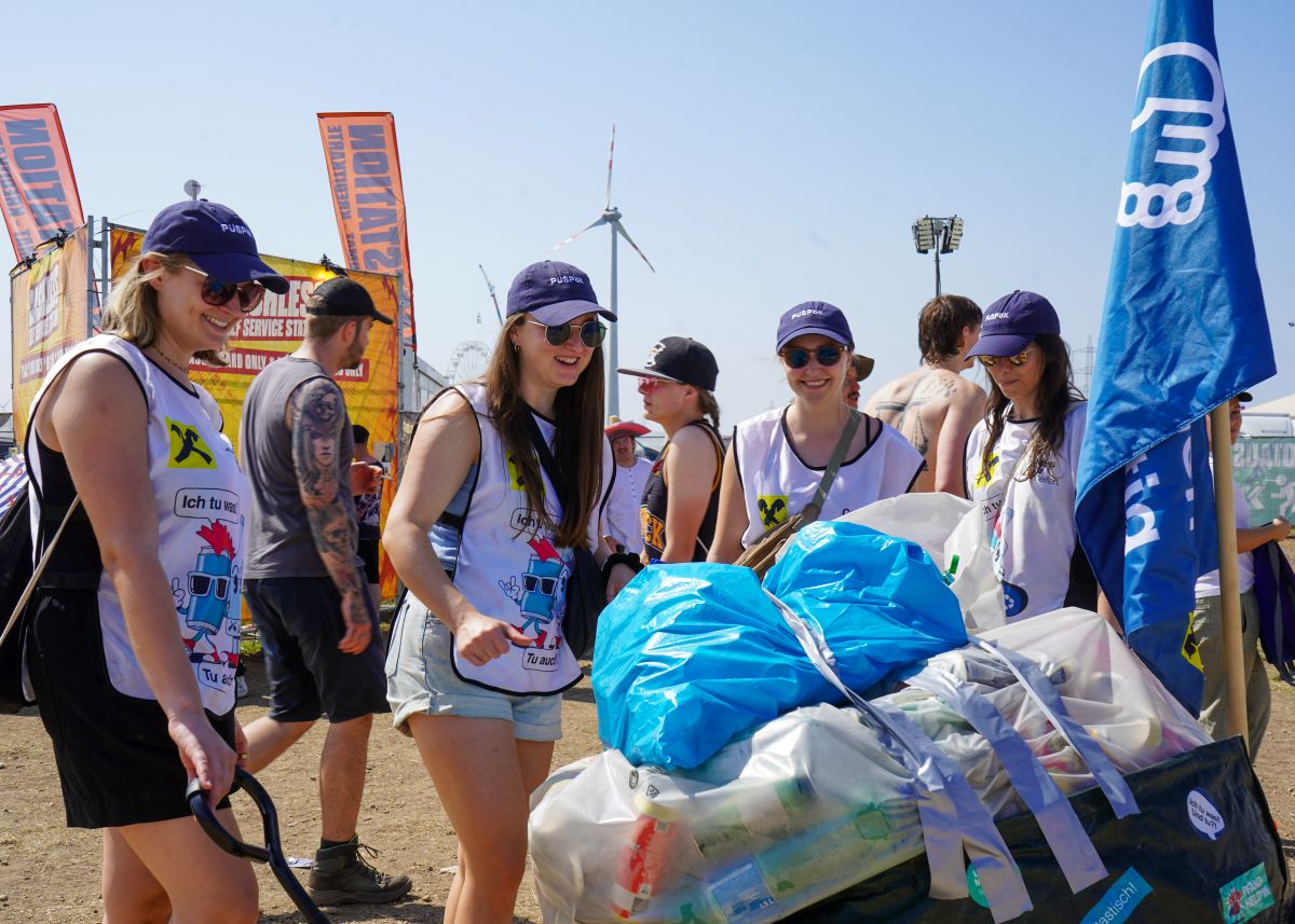 Vier junge Frauen mit Baseballkappen und Westen sammeln Pfandgebinde auf einem Festivalgelände, im Hintergrund stehen weitere Festivalbesucher unter blauem Himmel.