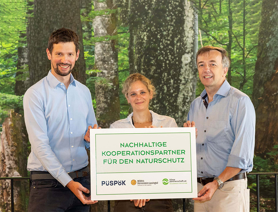 Drei Personen, zwei Männer und eine Frau, halten gemeinsam ein Schild mit der Aufschrift 'Nachhaltige Kooperationspartner für den Naturschutz' vor einem Hintergrund mit Bäumen. Auf dem Schild sind auch die Logos von Püspök, Wildnis Dürrenstein-Lassingtal und Klima Partnerschaft.at sichtbar.
