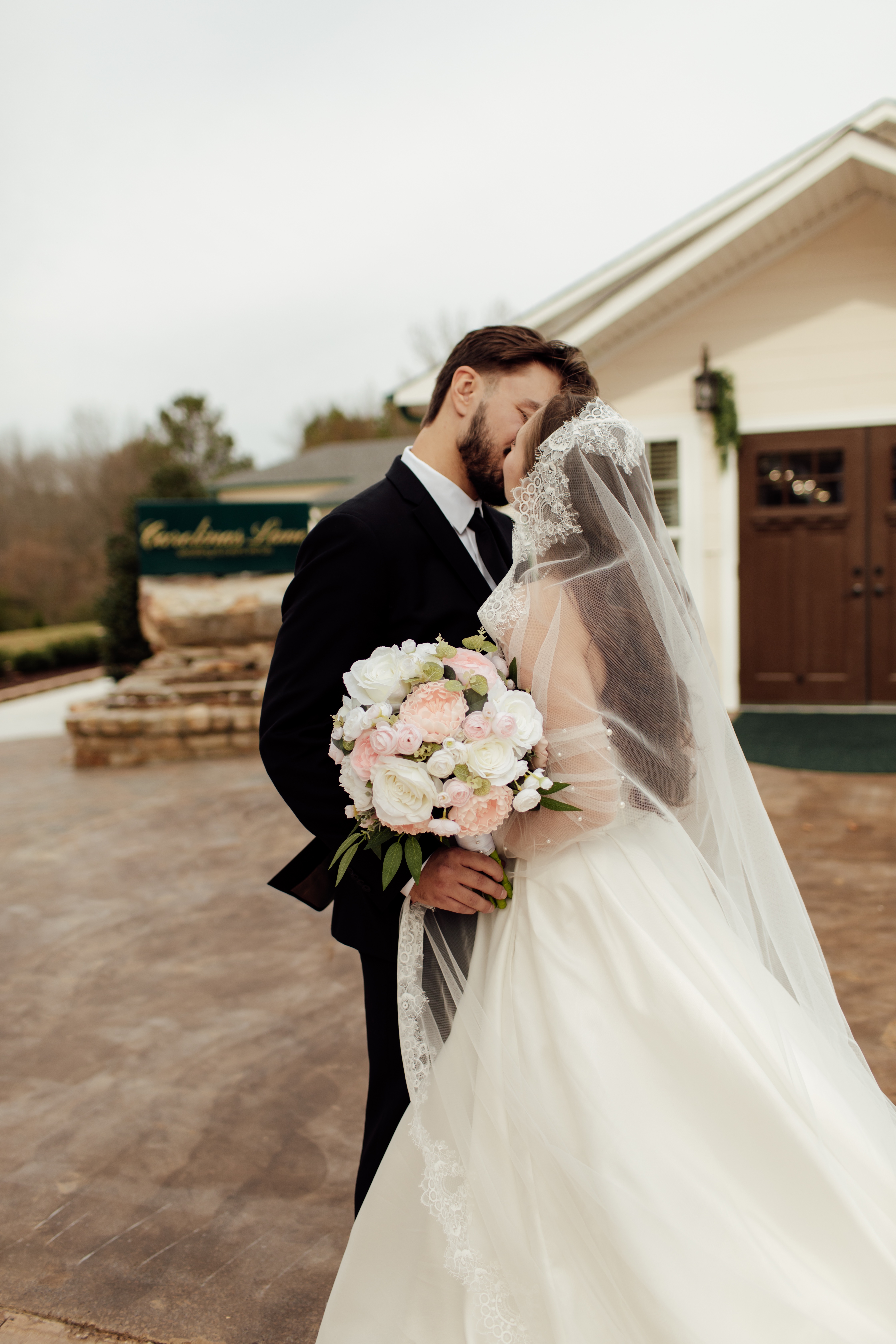 Bride and groom embracing and kissing outside a wedding venue, bride holding pink and white bouquet.